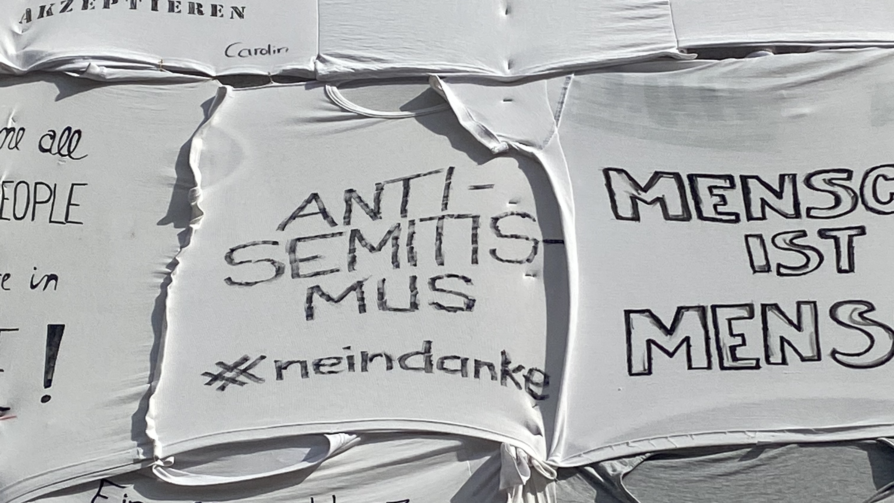 Collage aus weißen T-Shirts, welche per Hand in schwarzem Marker dekoriert sind. Auf dem sichtbarsten T-Shirt steht: "ANTISEMITISMUS #neindanke." Auf anderen: "Rote Karte gegen Diskriminierung, Rassismus und Antisemitismus." "Mensch ist Mensch." "Human Rights."