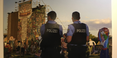 Photo of the removal of the controversial banner "People's Justice." The banner, half disassembled, is visible in the background while in the foreground, two police officers stand with their backs toward the camera.